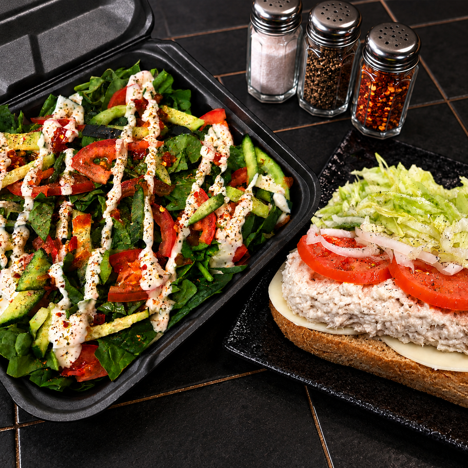 Fresh garden salad with creamy dressing alongside a tuna salad sandwich topped with lettuce and tomato, served with seasonings.