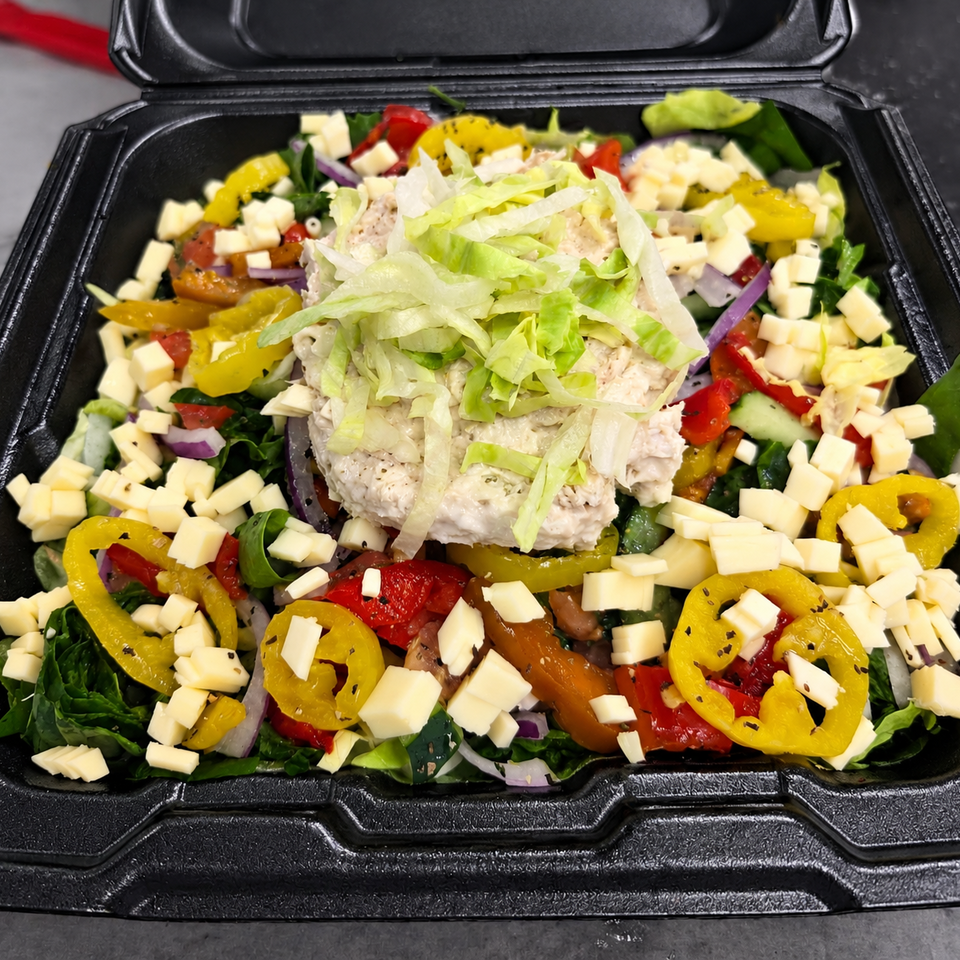 Chopped salad with creamy chicken salad, shredded lettuce, cheese, peppers, tomatoes, cucumbers, and onions in a black takeout container.