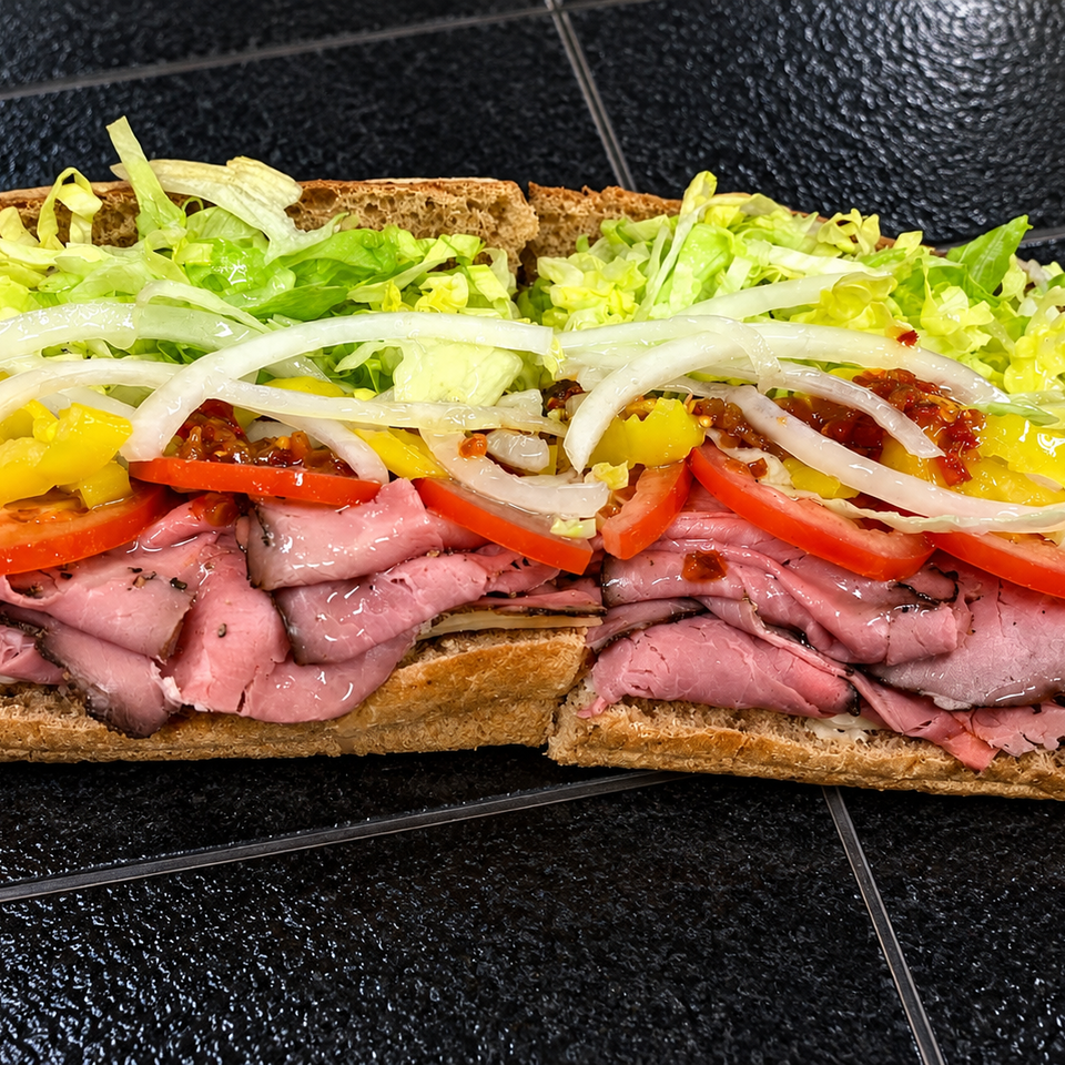 Overstuffed roast beef sandwich on toasted bread with lettuce, onions, tomatoes, peppers, and sauce on a black tiled surface.