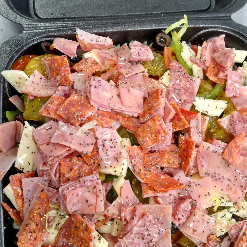 Close-up of a deli-style salad with chopped ham, pepperoni, salami, cheese, peppers, olives, and Italian dressing in a black container.